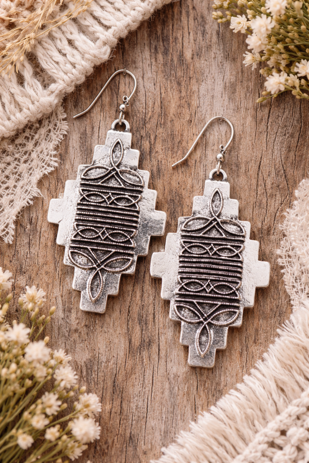 Pair of silver earrings on a wooden surface with floral and fabric accents