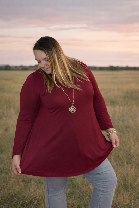 Woman wearing a red long-sleeve top and light blue jeans standing in a field at sunset.