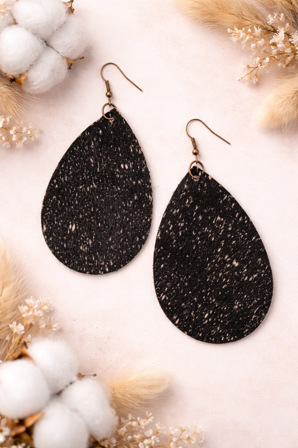 Black fur teardrop earrings on a soft white background with cotton and dried flowers.