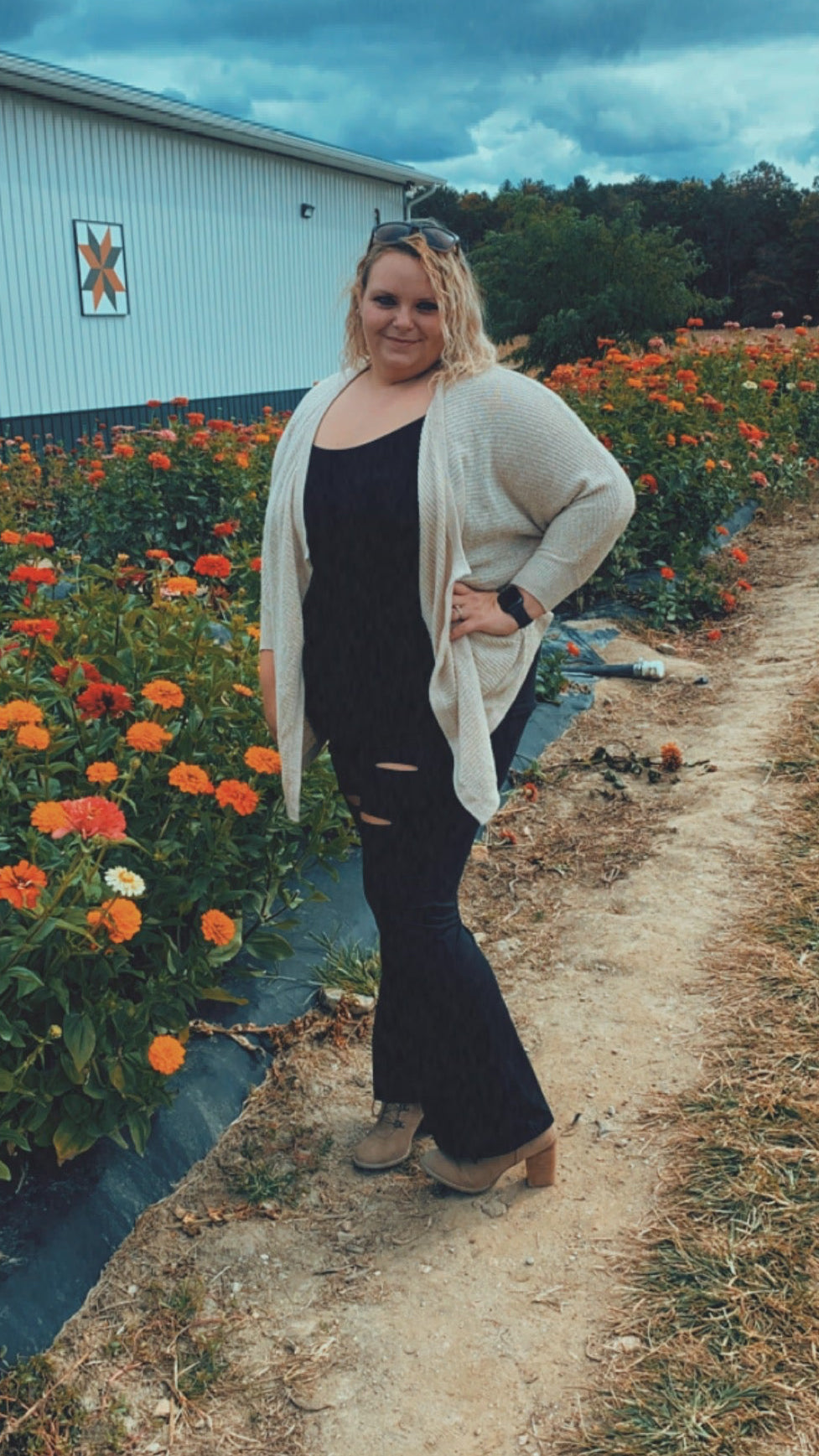 Woman standing on a dirt path with rows of orange flowers and a barn in the background.