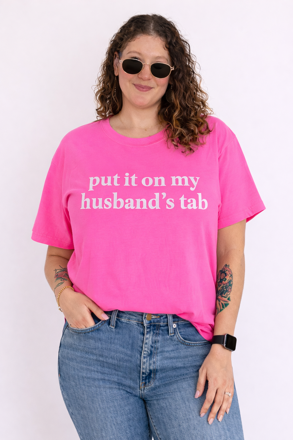 A woman wearing a hot pink shirt that says "put it on my husband's tab" with jeans on a white background. 