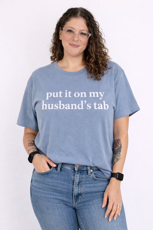 Person wearing a blue t-shirt with text 'put it on my husband's tab' on a white background