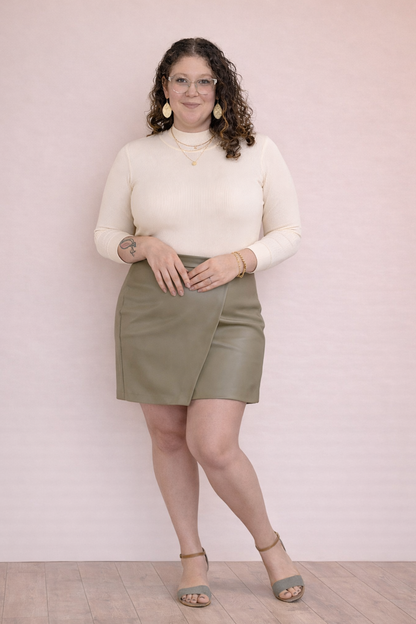 Woman wearing a leather olive skort and white shirt on a pale pink background. 