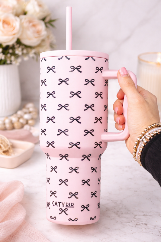 Pink mug with black bow pattern held by a hand, with a blurred background of flowers and a glass.