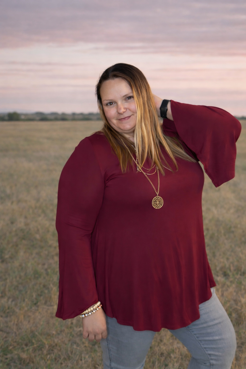 Woman in a red blouse standing in an open field with a sunset sky.