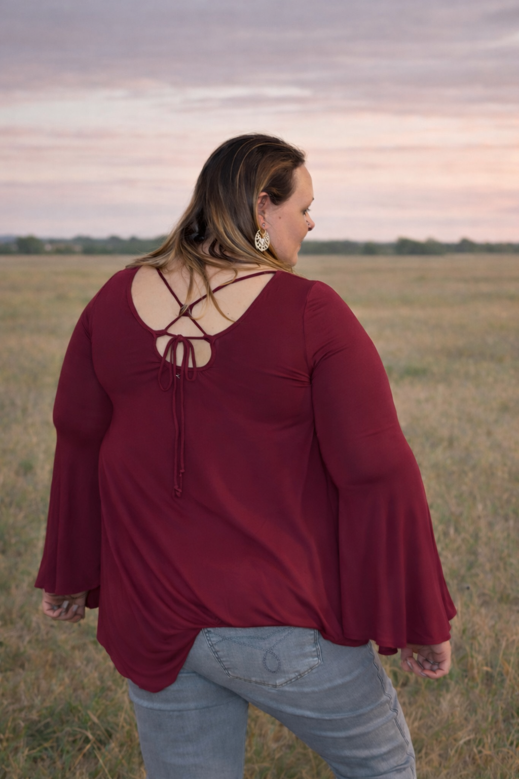 Woman wearing a burgundy blouse with a unique back design in an open field.