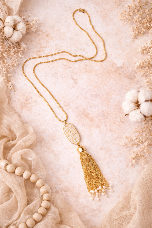 Gold necklace with tassel on a textured beige surface with cotton flowers